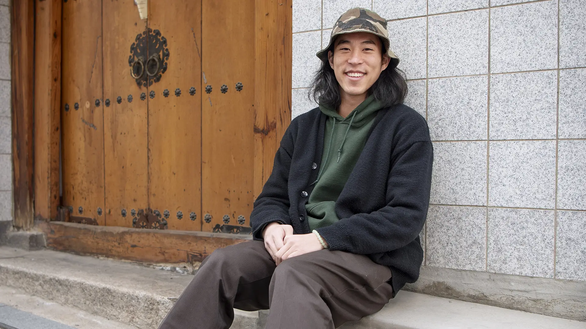 Korean man sitting down the street wearing a layered earth-tone cardigan and quiet luxury menswear.